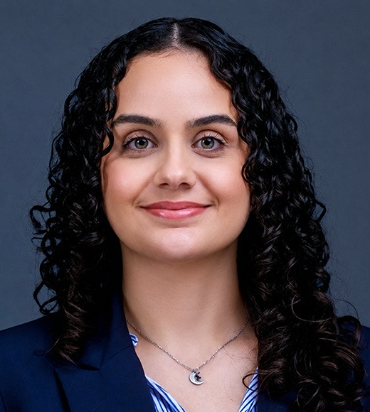 Headshot photograph of smiling woman with dark curly hair, navy blazer, gray background